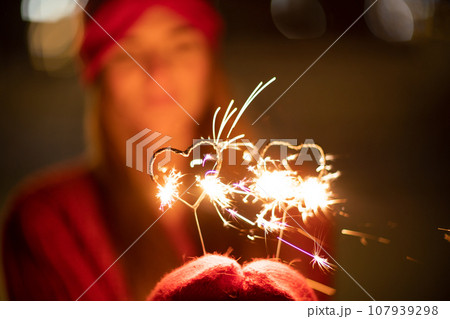 Woman holding sparkler night while celebrating Christmas outside. Dressed in a fur coat and a red headband. Blurred christmas decorations in the background. Selective focus 107939298