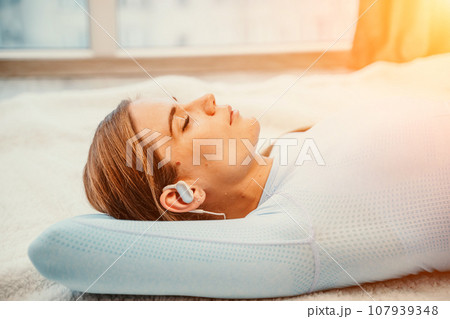 Side view portrait of relaxed woman listening to music with headphones lying on carpet at home. She is dressed in a blue tracksuit. Side view portrait of relaxed woman listening to music with headphones lying on carpet at home. She is dressed in a blue tracksuit. 107939348