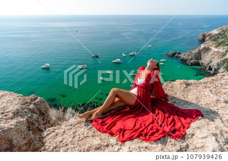 Woman red dress sea. Happy woman in a red dress and white bikini sitting on a rocky outcrop, gazing out at the sea with boats and yachts in the background. Woman red dress sea. Happy woman in a red dress and white bikini sitting on a rocky outcrop, gazing out at the sea with boats and yachts in the background. 107939426