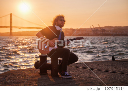 Hipster street musician in black playing electric guitar in the street on sunset on embankment with 25th of April bridge in background. Lisbon, Portugal Hipster street musician in black playing electric guitar in the street on sunset on embankment with 25th of April bridge in background. Lisbon, Portugal 107945114