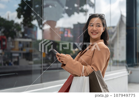 An attractive Asian woman holding her smartphone and shopping bags, enjoying her shopping day. 107949775