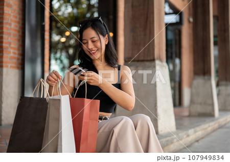 A gorgeous Asian woman is taking pictures of her shopping bags while resting on the stairs outside. 107949834