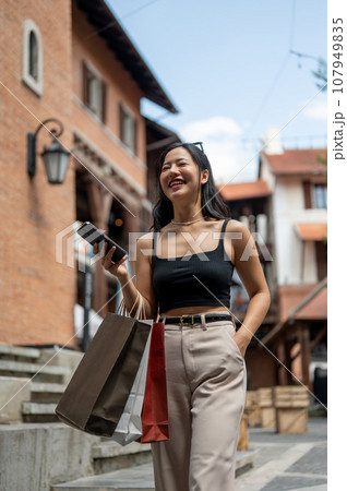 An attractive woman enjoys shopping in the city, walking while holding her phone and shopping bags. 107949835