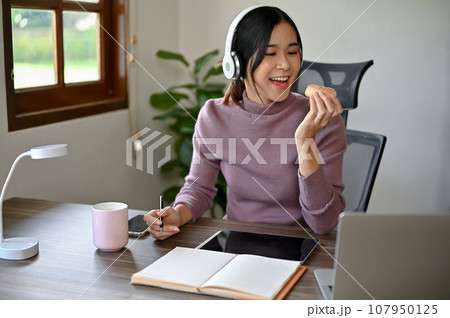 Beautiful Asian girl eating a donut while studying at her study table in her house 107950125