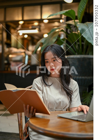 A beautiful Asian woman focuses on reading a book while working remotely at a coffee shop. 107950332