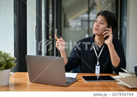 A smart Asian male office worker is on the phone with a business supplier while sitting at his desk A smart Asian male office worker is on the phone with a business supplier while sitting at his desk 107950704