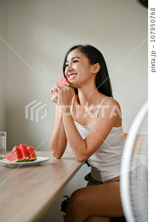A smiling Asian woman enjoys her fresh watermelons while sitting in front of a fan A smiling Asian woman enjoys her fresh watermelons while sitting in front of a fan 107950768