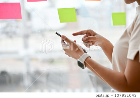 Close-up image of a woman using her smartphone while standing by the window in her office. 107951045