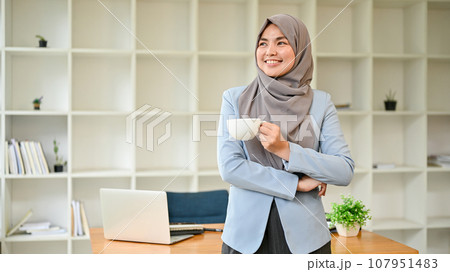 A confident Asian Muslim businesswoman stands in her office with a coffee cup in her hands. A confident Asian Muslim businesswoman stands in her office with a coffee cup in her hands. 107951483