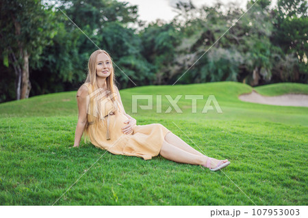 Happy young pregnant model sitting on grass lawn and looking at setting sun in park. Smiling future mom expecting baby. Maternity concept. Copy space Happy young pregnant model sitting on grass lawn and looking at setting sun in park. Smiling future mom expecting baby. Maternity concept. Copy space 107953003