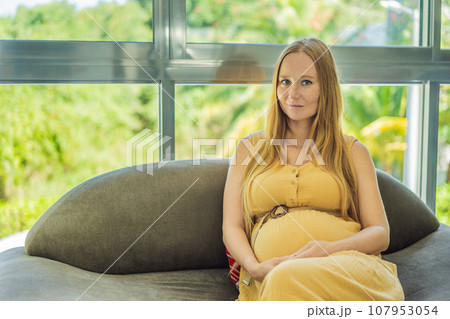Beautiful pregnant woman sitting at sofa and keeping hand on belly Beautiful pregnant woman sitting at sofa and keeping hand on belly 107953054
