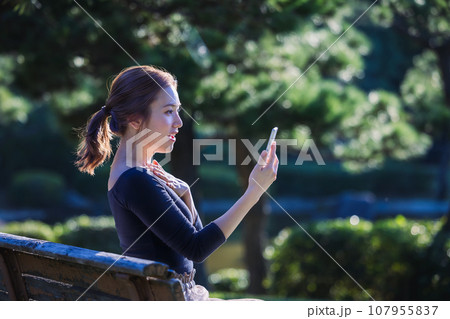 休日に公園でスマートフォンを見る若い女性 休日に公園でスマートフォンを見る若い女性 107955837