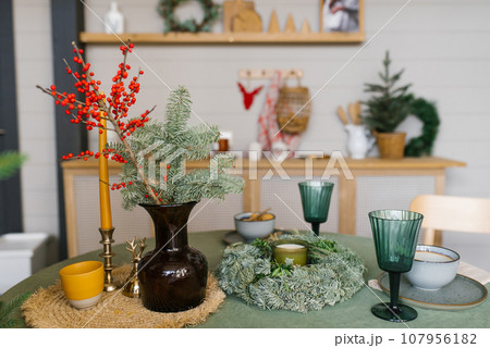 Festive table set in the kitchen for Christmas and New Year. Spruce branches in a vase, glass glasses, a candle, a wreath on the table 107956182