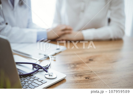 Stethoscope and laptop computer are lying on the wooden table while doctor's hands reassuring a patient at the background. Medicine concept Stethoscope and laptop computer are lying on the wooden table while doctor's hands reassuring a patient at the background. Medicine concept 107959589