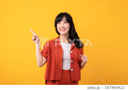 Portrait young beautiful asian woman happy smile dressed in orange clothes showing pointing finger hand gesture to free space isolated on yellow studio background. Attention to nice product 107960422