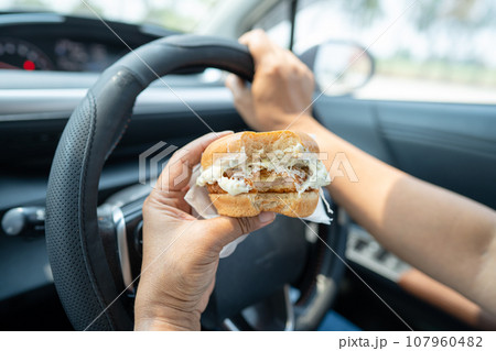 Asian lady holding hamburger to eat in car, dangerous and risk an accident. 107960482