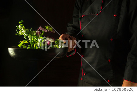 The cook prepares fresh vegetables in a frying pan. Grande cuisine on dark background. Free advertising space. Frying pan with flying vegetables in the chef hand. The cook prepares fresh vegetables in a frying pan. Grande cuisine on dark background. Free advertising space. Frying pan with flying vegetables in the chef hand. 107960920