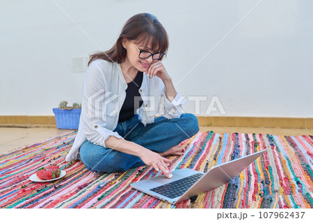 Happy middle-aged woman sitting on floor at home using laptop 107962437