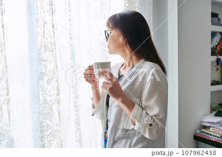 Serious middle-aged woman looking out window, with cup in hands Serious middle-aged woman looking out window, with cup in hands 107962438