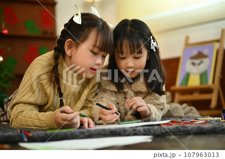 Cute little girls lying on floor in living room decorated for Christmas and making Christmas greeting card Cute little girls lying on floor in living room decorated for Christmas and making Christmas greeting card 107963013