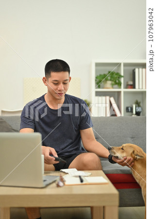 Smiling asian man in casual clothes playing with lovely dog while sitting on sofa living room Smiling asian man in casual clothes playing with lovely dog while sitting on sofa living room 107963042