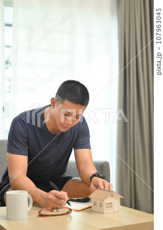 Concentrated millennial man hand writing notes for planning and calculating money for buy house or estates Concentrated millennial man hand writing notes for planning and calculating money for buy house or estates 107963045