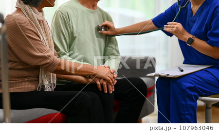 Female doctor using stethoscope checking heart and lungs of senior man during home visit. Elderly healthcare concept Female doctor using stethoscope checking heart and lungs of senior man during home visit. Elderly healthcare concept 107963046