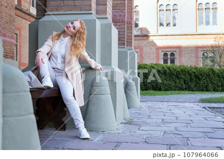 Woman waiting someone and rests outside leaning on the wall. Young woman in light clothes sits on bench near column and looks sideways. Woman waiting someone and rests outside leaning on the wall. Young woman in light clothes sits on bench near column and looks sideways. 107964066