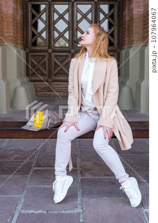 Smiling young woman in white clothes sits on bench with bouquet of flowers on historic building background. Vertical frame. 107964067