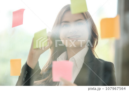 Beautiful female employee write down on colorful sticky notes manage list, concentrated biracial woman work on startup brainstorm collaborate plan on stickers on glass wall. 107967564