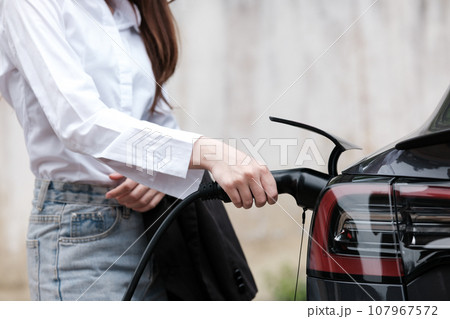 Young woman traveling by electric car having stop at charging station standing plugging charger close-up. Young woman traveling by electric car having stop at charging station standing plugging charger close-up. 107967572