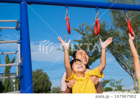Cute little girl hanging bars in the playground. Happy little Asian child having fun on playground, climbing bars in park. Little girl playing in outdoor playground with mother. 107967902