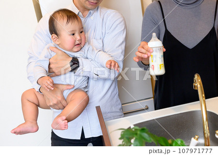 父親に抱っこしてもらい赤ちゃんに冷やしたミルクを飲ませる母親　あやす　乳幼児　イクメン　0歳乳幼児 107971777