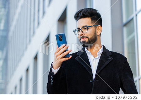 Stylish business man using smartphone typing text answering messages chatting online in city street Stylish business man using smartphone typing text answering messages chatting online in city street 107975505