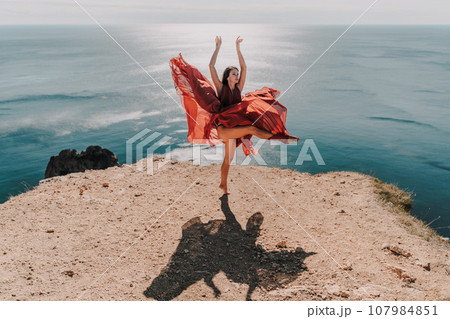 Woman red dress sea. Female dancer posing on a rocky outcrop high above the sea. Girl on the nature on blue sky background. Fashion photo. Woman red dress sea. Female dancer posing on a rocky outcrop high above the sea. Girl on the nature on blue sky background. Fashion photo. 107984851