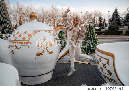 Woman Christmas Square. She stands near a large white cup, dressed in a light suit. With trees decorated with Christmas tinsel in the background 107984934