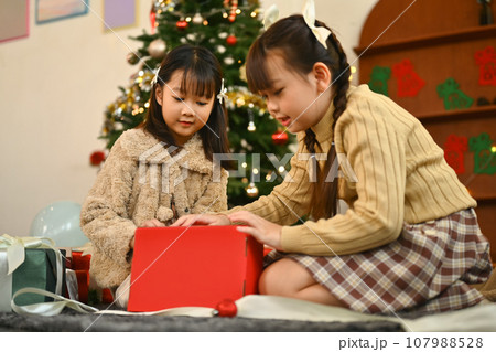 Two adorable girls ecstatically opening Christmas gift box near decorated Christmas tree in a cozy living room 107988528
