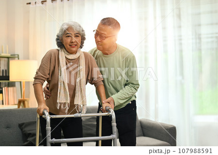 Caring senior man helping his wife walking with walker at home. Elderly healthcare concept Caring senior man helping his wife walking with walker at home. Elderly healthcare concept 107988591