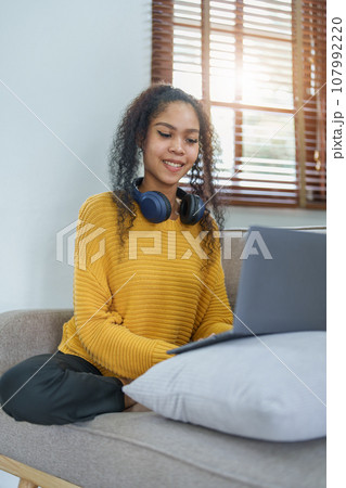Portrait of an African American sitting on the sofa wearing on-ear headphones and using a computer at home. Portrait of an African American sitting on the sofa wearing on-ear headphones and using a computer at home. 107992220