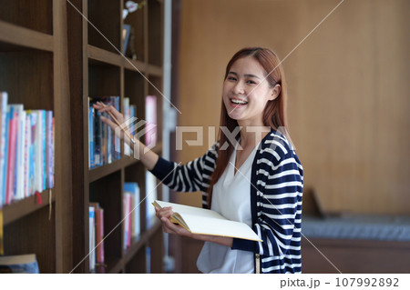 Portrait of a young Asian woman showing joy as she searches for knowledge in the library. Portrait of a young Asian woman showing joy as she searches for knowledge in the library. 107992892