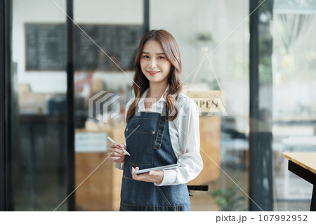 open a small business Portrait of happy Asian woman in apron holding tablet in front of cafe open a small business Portrait of happy Asian woman in apron holding tablet in front of cafe 107992952