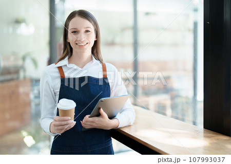 Starting and opening a small business, a young Asian woman showing a smiling face in an apron standing in front of a coffee shop bar counter. Business Owner, Restaurant, Barista, Cafe, Online SME. 107993037