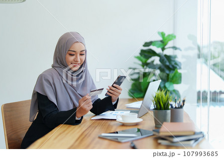Beautiful Muslim woman shopping online using her phone and credit card. Beautiful Muslim woman shopping online using her phone and credit card. 107993685