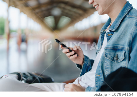 Asian teenage male traveler using her smart phone mobile while waiting for a train at a station Asian teenage male traveler using her smart phone mobile while waiting for a train at a station 107994154
