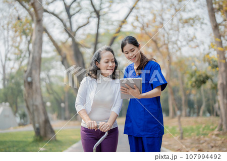 Patient care,, young Asian female nurse caring for with a cane and doing leg and knee exercises using a digital tablet as a teaching tool during a walk in the park parks 107994492