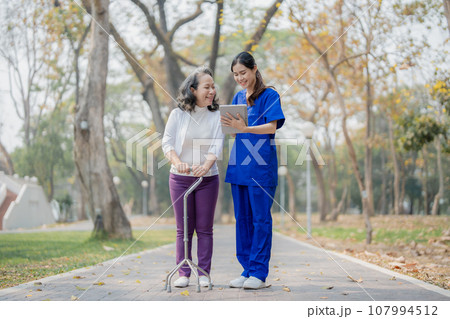 Patient care,, young Asian female nurse caring for with a cane and doing leg and knee exercises using a digital tablet as a teaching tool during a walk in the park parks Patient care,, young Asian female nurse caring for with a cane and doing leg and knee exercises using a digital tablet as a teaching tool during a walk in the park parks 107994512