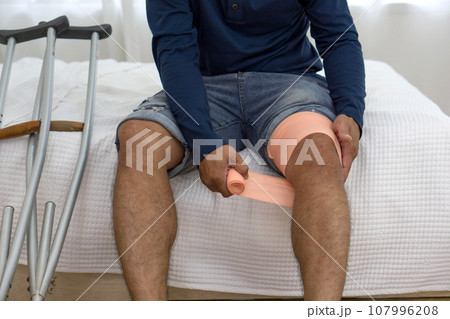 A man with a broken knee sitting on his bed. He leans on crutches, signalling recovery from injury. His bedroom serves as a backdrop, tinged with hints of helplessness yet enduring determination. 107996208