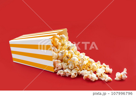 Tasty cheese popcorn falling out of a yellow striped carton bucket, isolated on red background Tasty cheese popcorn falling out of a yellow striped carton bucket, isolated on red background 107996796