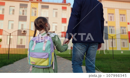 The father holds the hand of a first-grader with a backpack. Happy family. The father accompanies the child to school. The girl hugs her dad. My daughter is a girl with a backpack. Keith goes to the 107998345