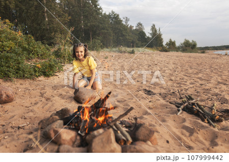happy smiling cheerful girl 6 years old sits sitting on beach near forest opposite fire and enjoys in summer day, picnic, vacation 107999442